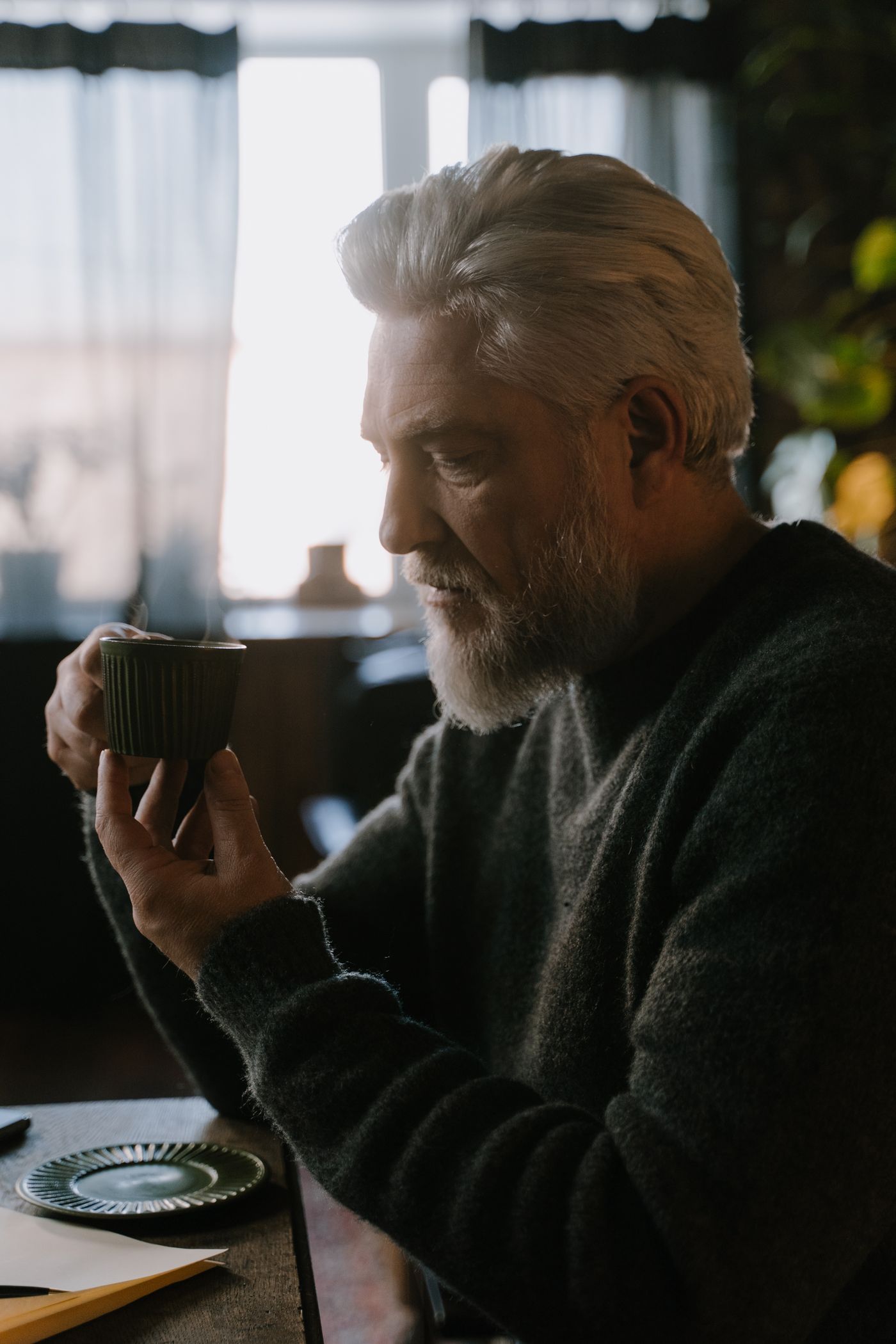 A grey-bearded man with a cup of coffee by the window